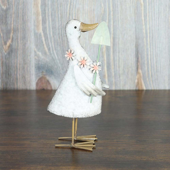 Goldbach – Ente mit Spaten steht auf einer Holzfläche, hält eine grüne Schaufel und trägt einen Kranz aus rosa Blumen.