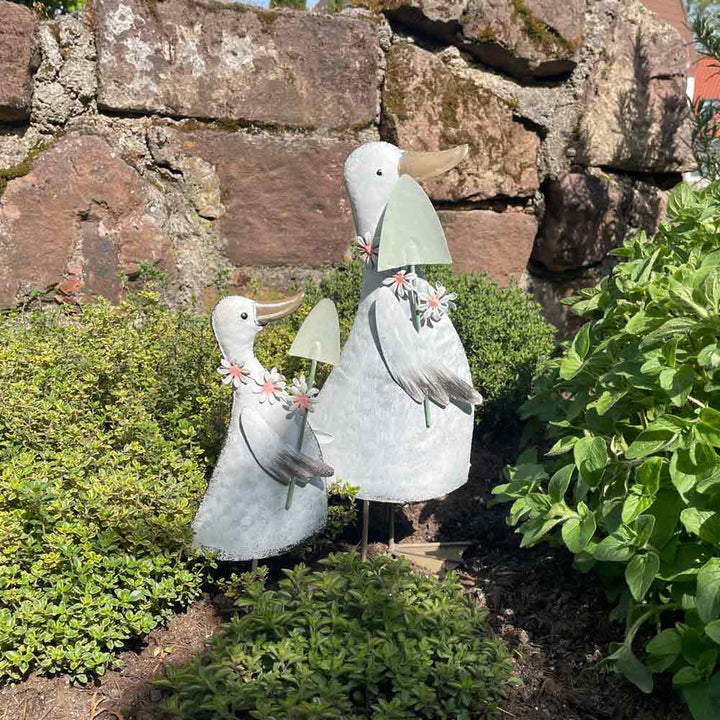 Zwei Goldbach-Ente-mit-Spaten-Statuen mit Blumensträußen stehen im Grünen neben einer rustikalen Steinmauer.