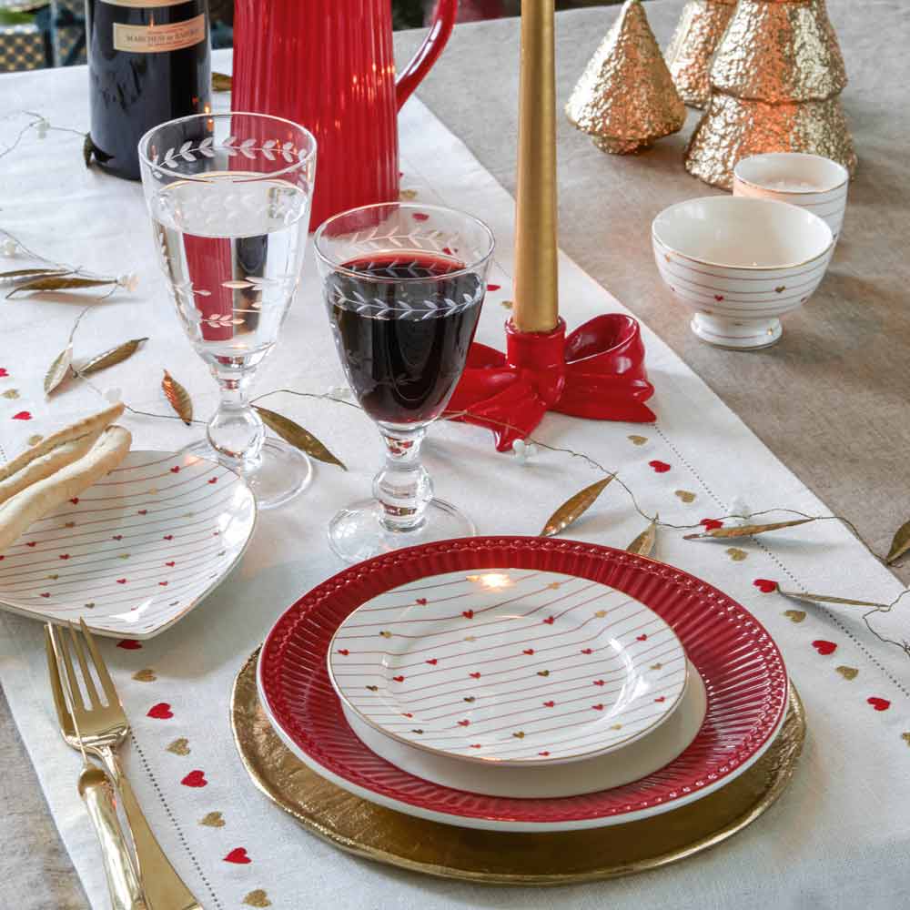 Für ein festliches Essen gedeckter Tisch mit rot-goldenen Tellern, einem GreenGate - Grace Mini Latte Cup, Weingläsern, einer Kerze und dekorativen Bäumen auf einem weiß-silbernen Tischläufer.