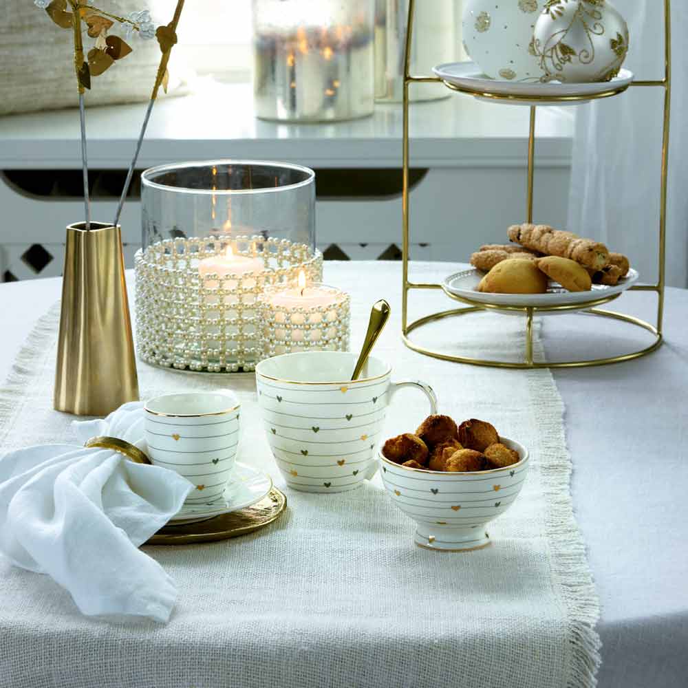 Ein gedeckter Tisch mit einer weißen Tischdecke, Kerzen, einer GreenGate - Grace Mini Latte Cup, einer Tasse, einem Teller mit Snacks, einer Serviette und einem Etagere mit Keksen.