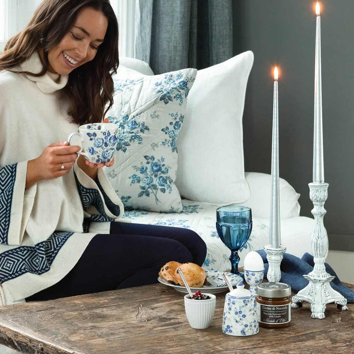 Eine Frau sitzt auf einem Sofa und hält eine Tasse mit Blumenmuster in der Hand. Neben einem Holztisch steht ein großer GreenGate-Kerzenständer Emaille, ein Ei in einer Tasse, Marmelade, ein Scone, ein blaues Glas und andere Gegenstände.