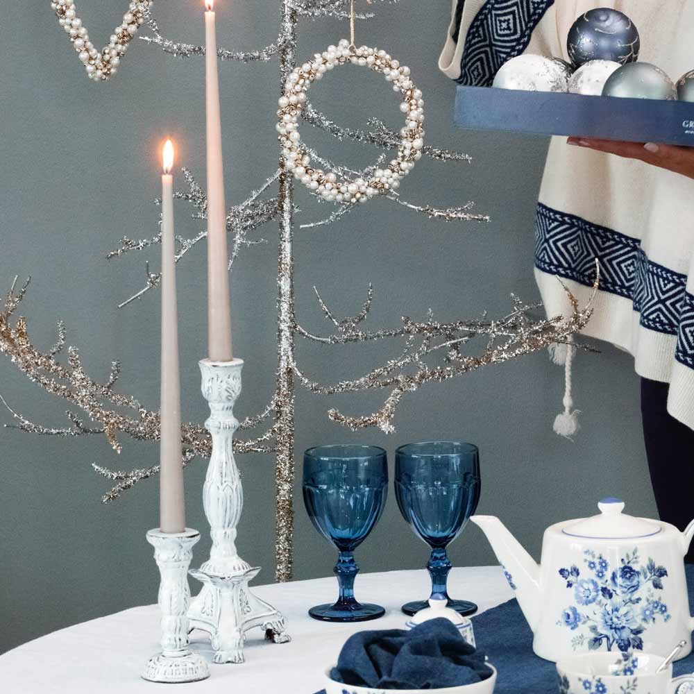 Zwei beleuchtete GreenGate - Kerzenständer Emaille groß in weißen Haltern, ein Kranz, zwei blaue Weingläser, eine weiße Teekanne mit blauen Blumen und eine Person, die eine Schachtel mit Weihnachtsschmuck hält, arrangiert auf einem Tisch mit einer weißen Tischdecke.