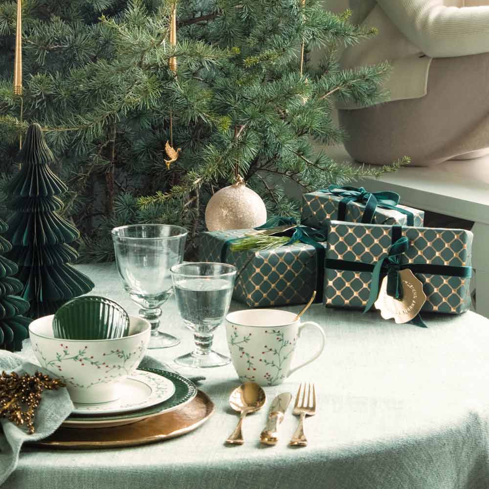 Ein für Weihnachten gedeckter Tisch mit GreenGate - Lucie Xmas Tannen Teller weiß, Tassen, Besteck und Gläsern. Im Hintergrund steht ein Weihnachtsbaum mit Ornamenten und verpackten Geschenken.