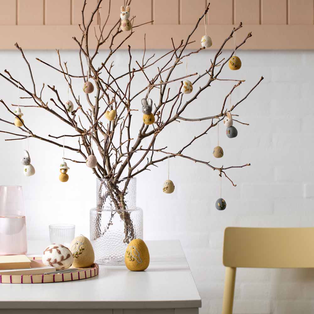 Ein minimalistisches Oster-Display mit einer Vase mit kahlen Zweigen, geschmückt mit dem 3er-Set „Gry & Sif – Anhänger Ei mit Blättern Filz“, begleitet von weiteren Eiern, die auf dem Tisch liegen.