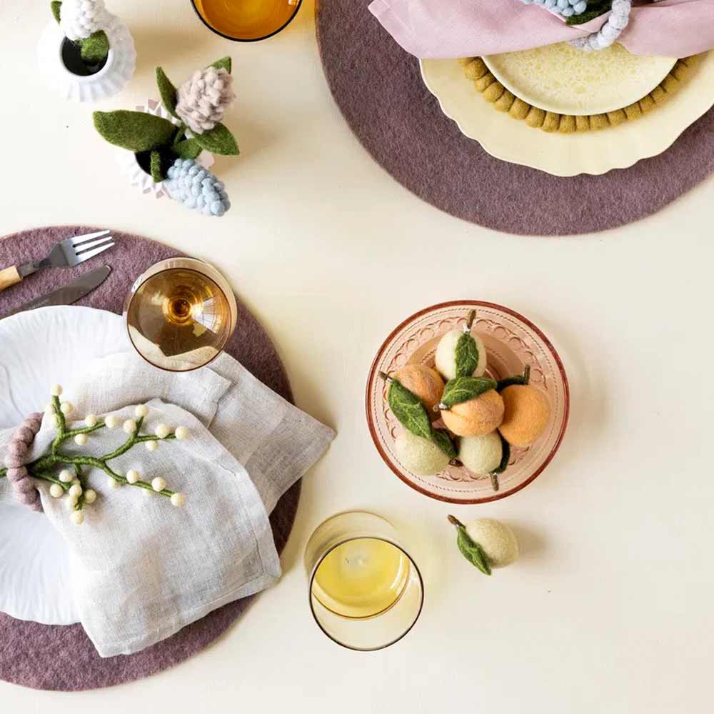 Ein gedeckter Tisch mit Tellern, Servietten, Weingläsern und dekorativem Obst von Gry & Sif - Zitrone Filz auf Tischsets; außerdem sind Utensilien und eine kleine Vase mit Filzblumen zu sehen.