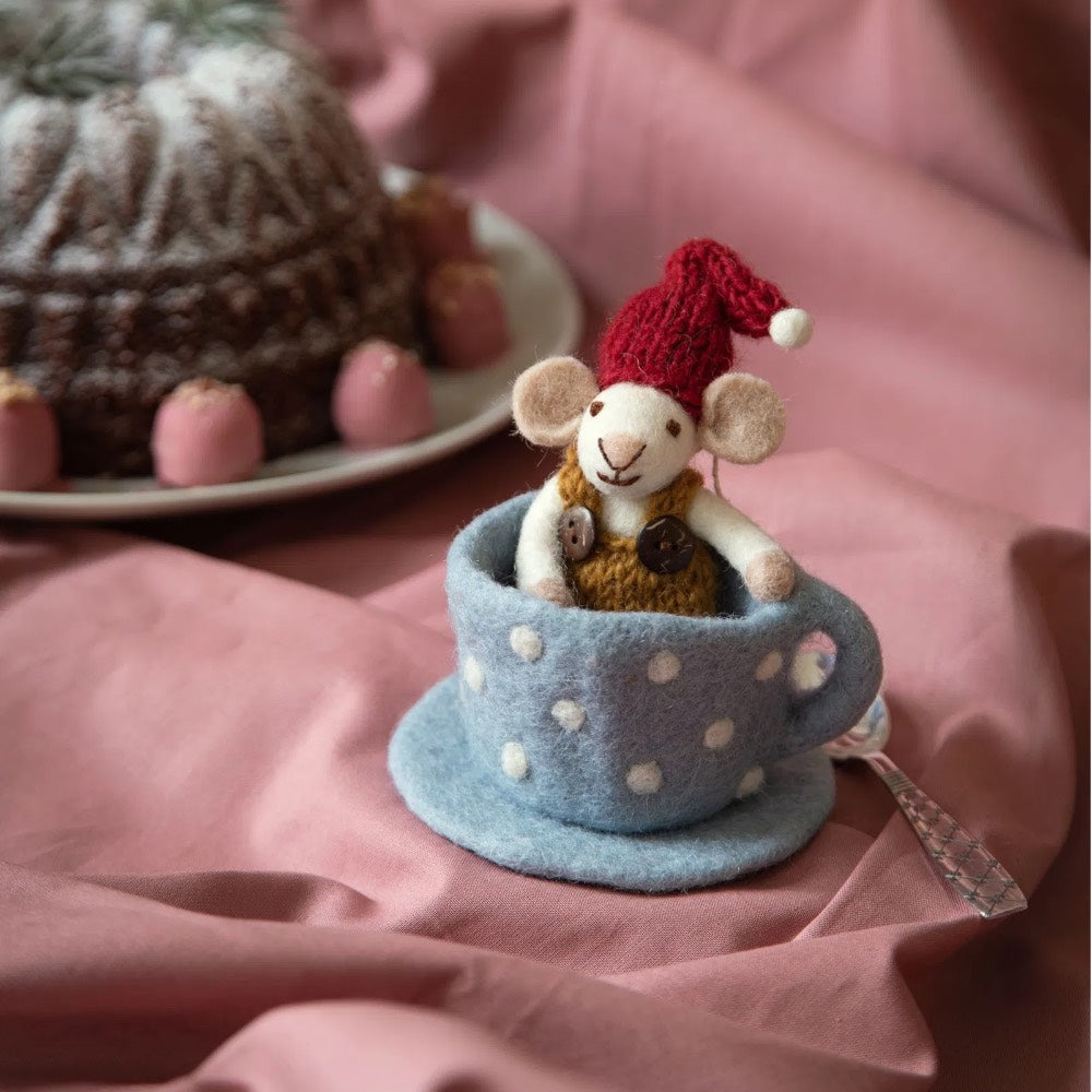 Die Gry & Sif - Maus Filz in blauer Tasse zeigt eine kleine Filzmaus mit rotem Hut, die in einer blau gepunkteten Teetasse auf rosa Stoff sitzt, umgeben von Kuchen und rosa Leckereien im Hintergrund.