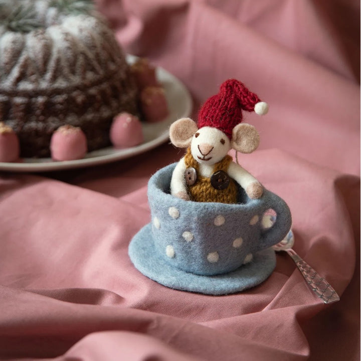 Die Gry & Sif - Maus Filz in blauer Tasse zeigt eine kleine Filzmaus mit rotem Hut, die in einer blau gepunkteten Teetasse auf rosa Stoff sitzt, umgeben von Kuchen und rosa Leckereien im Hintergrund.