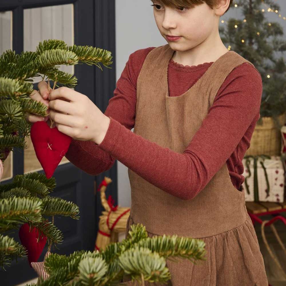 Ein Kind hängt das Ornament „Ib Laursen - Anhänger Herz Stoff My Nostalgic Christmas“ an einen Weihnachtsbaum. Es trägt ein braunes Kleid über einem roten Hemd. Im Hintergrund sind Geschenke und festliche Dekorationen zu sehen.