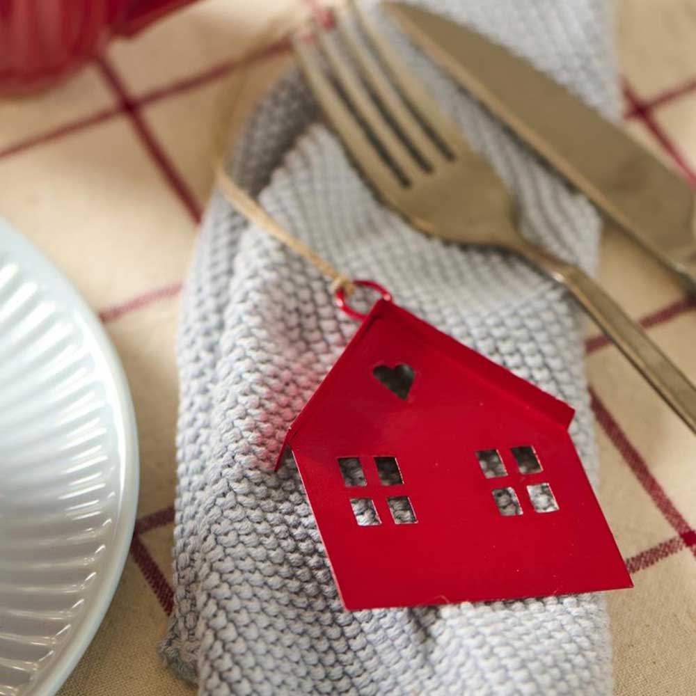 Der Ib Laursen - Anhänger Metallhaus rot, ein rotes hausförmiges Ornament mit ausgeschnittenen Fenstern und einem Herz, liegt auf einer gefalteten grauen Serviette neben Gabel und Messer auf einer karierten Tischdecke.