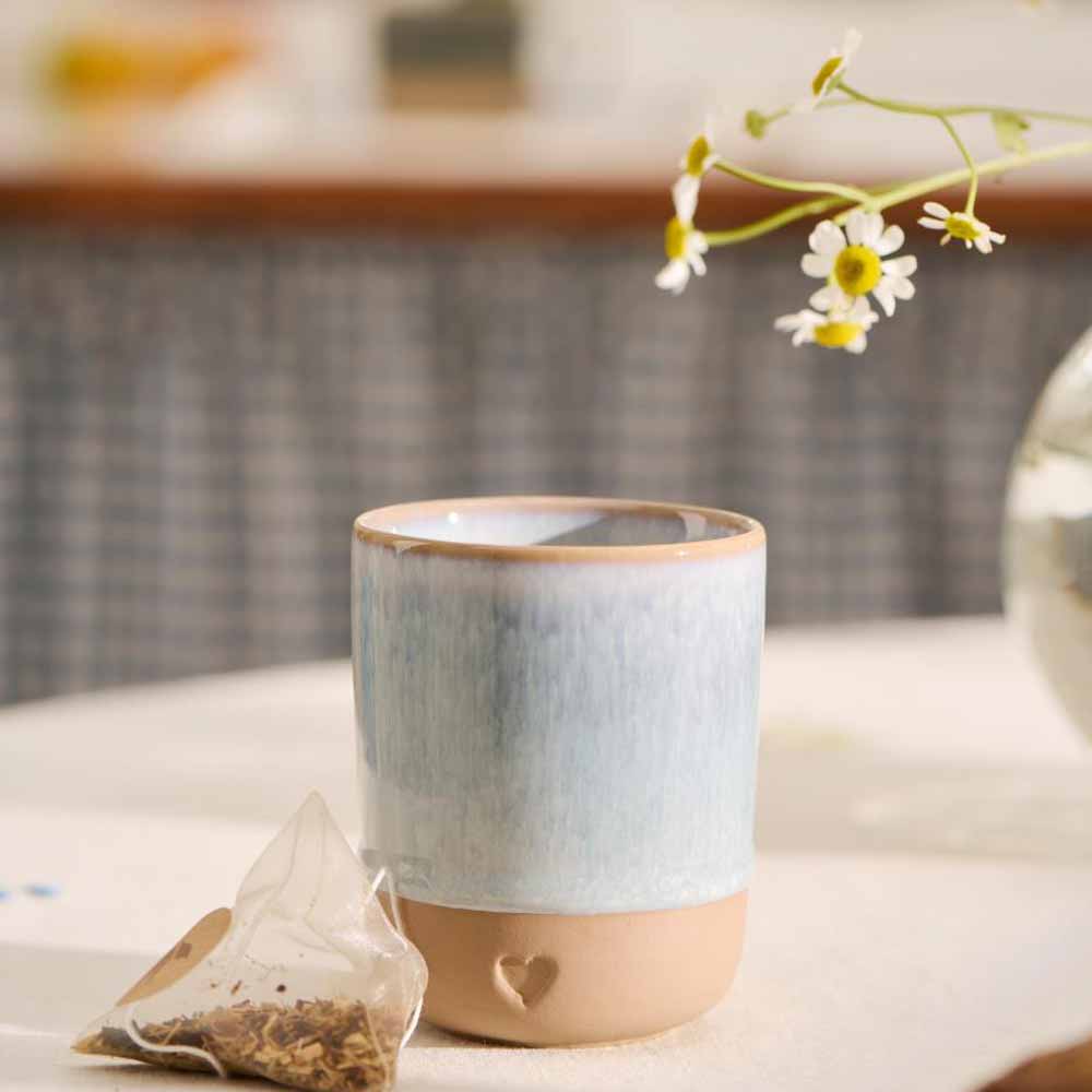 Der Ib Laursen - Becher Herz Ton Natur mit hellblauer Glasur steht neben einem pyramidenförmigen Teebeutel und einer kleinen Vase mit weißen Blumen auf dem Tisch.