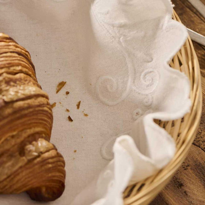 Ein teilweise gegessenes Croissant liegt in einem runden Weidenkorb, der mit einer Ib Laursen - Brotserviette rund weiß mit Stickerei ausgelegt ist, auf deren Stoff sichtbare Krümel verstreut sind.