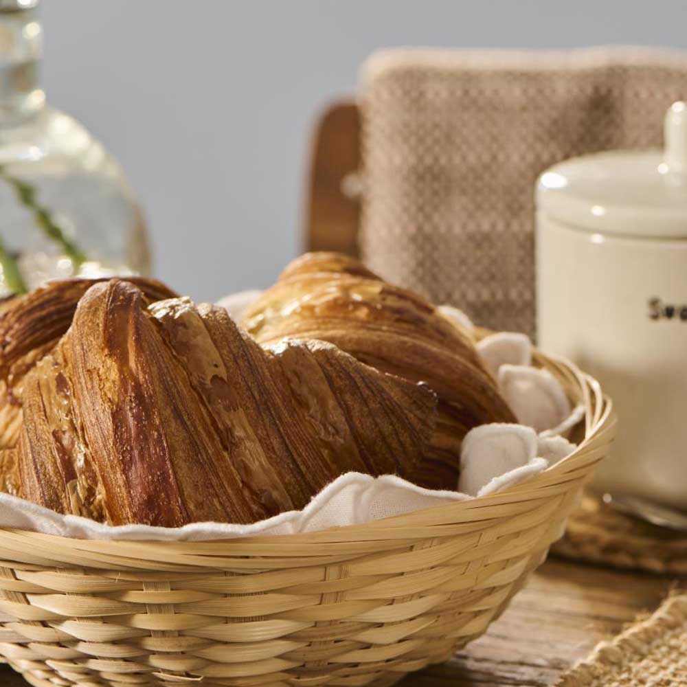 Ein Weidenkorb mit zwei Croissants, bedeckt mit einer runden weißen Brotserviette von Ib Laursen mit Stickerei, steht auf einem Holztisch neben einem Zuckerbehälter und einer Vase.