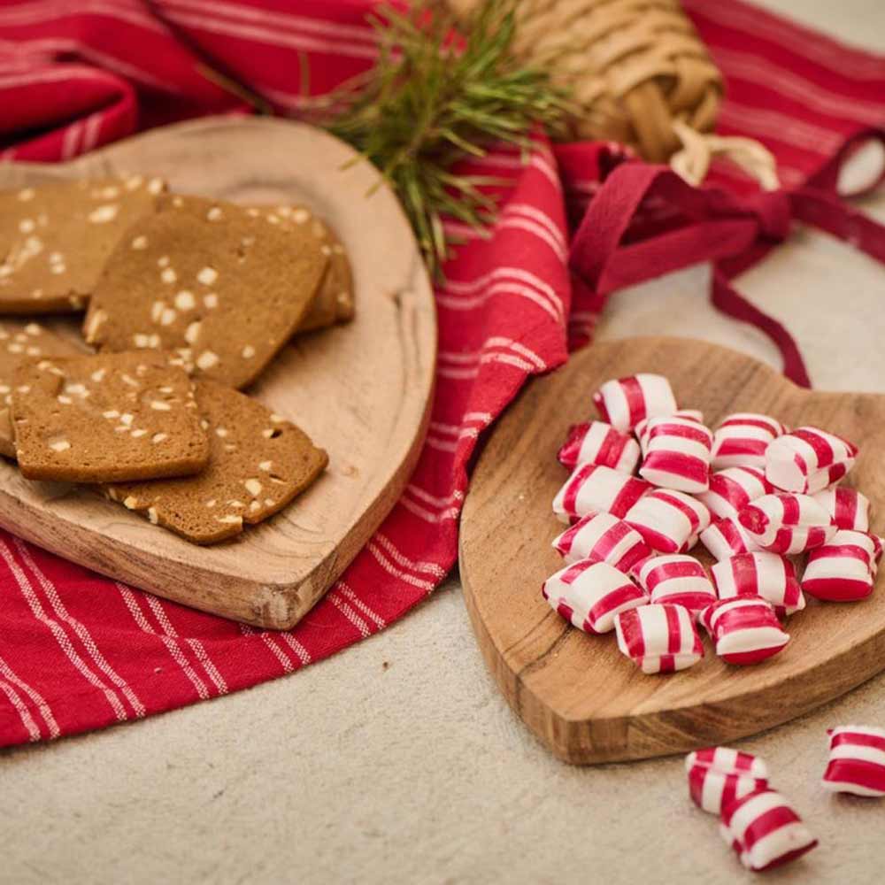 Holztabletts mit geschnittenen Lebkuchen und gestreiften rot-weißen Pfefferminzbonbons auf einem Ib Laursen Geschirrtuch Kaja in Rot mit dünnen naturfarbenen Streifen. Im Hintergrund sind Tannenzweige und eine Weidenglocke zu sehen.