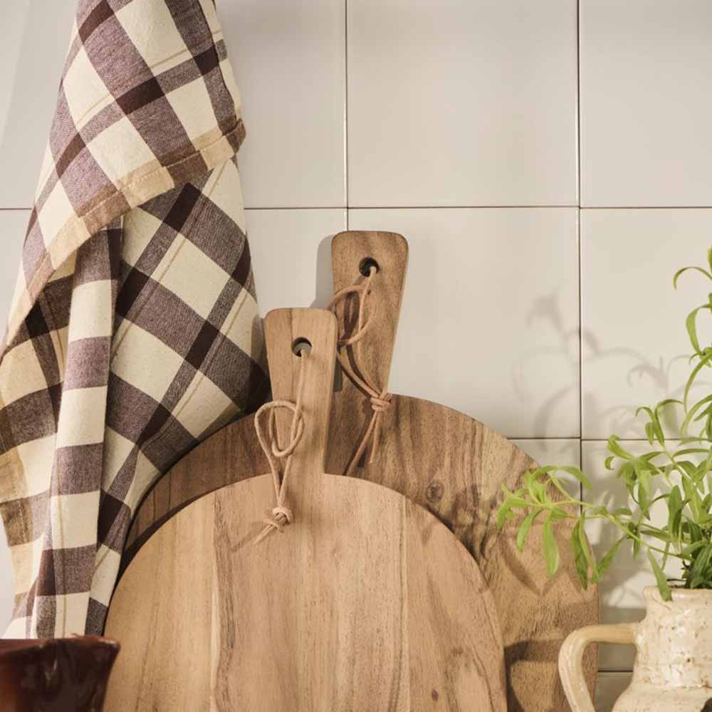 Zwei hölzerne Schneidebretter und ein Ib Laursen - Geschirrtuch Malthe natur mit dunkelbraunem Streifenmuster lehnen an einer gefliesten Wand, auf der rechten Seite ist eine grüne Pflanze teilweise sichtbar.