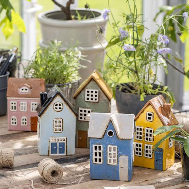 Eine Sammlung von Ib Laursen - Haus für Teelicht Nyhavn gerundete Dachfenster in verschiedenen Farben wird auf einem Holztisch ausgestellt. Topfpflanzen und Gartengeräte sind im Hintergrund zu sehen.