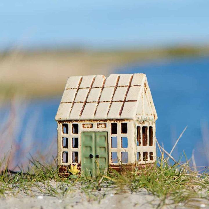 Das Ib Laursen - Haus für Teelicht Orangerie ist ein kleines Keramikhaus mit einer grünen Tür, das auf einer Wiese am Wasser steht, mit einer verschwommenen Landschaft im Hintergrund.