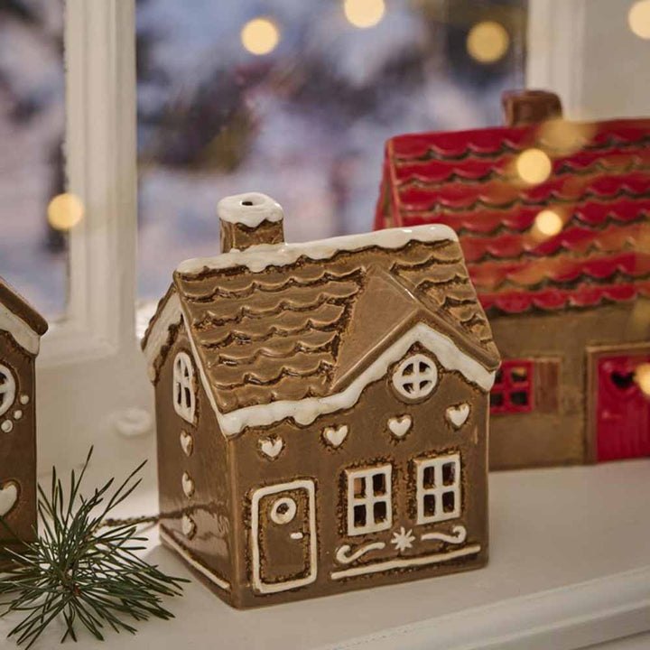 Eine viereckige Tür von Ib Laursen - Haus für Teelicht Stillenat Gingerbread, verziert mit aufwendigen weißen Glasurdetails, ruht auf einer Fensterbank, verbunden mit einem weiteren Haus im Hintergrund und sanft beleuchtet von warmen Lichterketten.