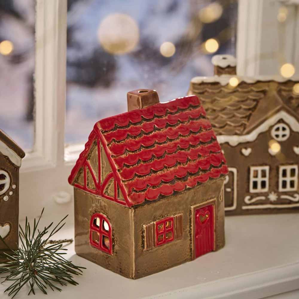 Das Ib Laursen Haus für Teelicht-Stillenat mit rotem Dach und roter Tür steht auf einer Fensterbank mit einer verschneiten Außenszene und Weihnachtsbeleuchtung im Hintergrund.