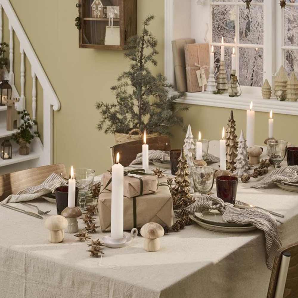 Ein festlich gedeckter Esstisch mit dem Ib Laursen Kerzenhalter Pilz mit matter Glasur für Stabkerzen, dazu ein verpacktes Geschenk, Holzpilze und Baumdeko. Der Hintergrund zeigt eine Treppe und ein Fenster, die eine verschneite Landschaft einrahmen.