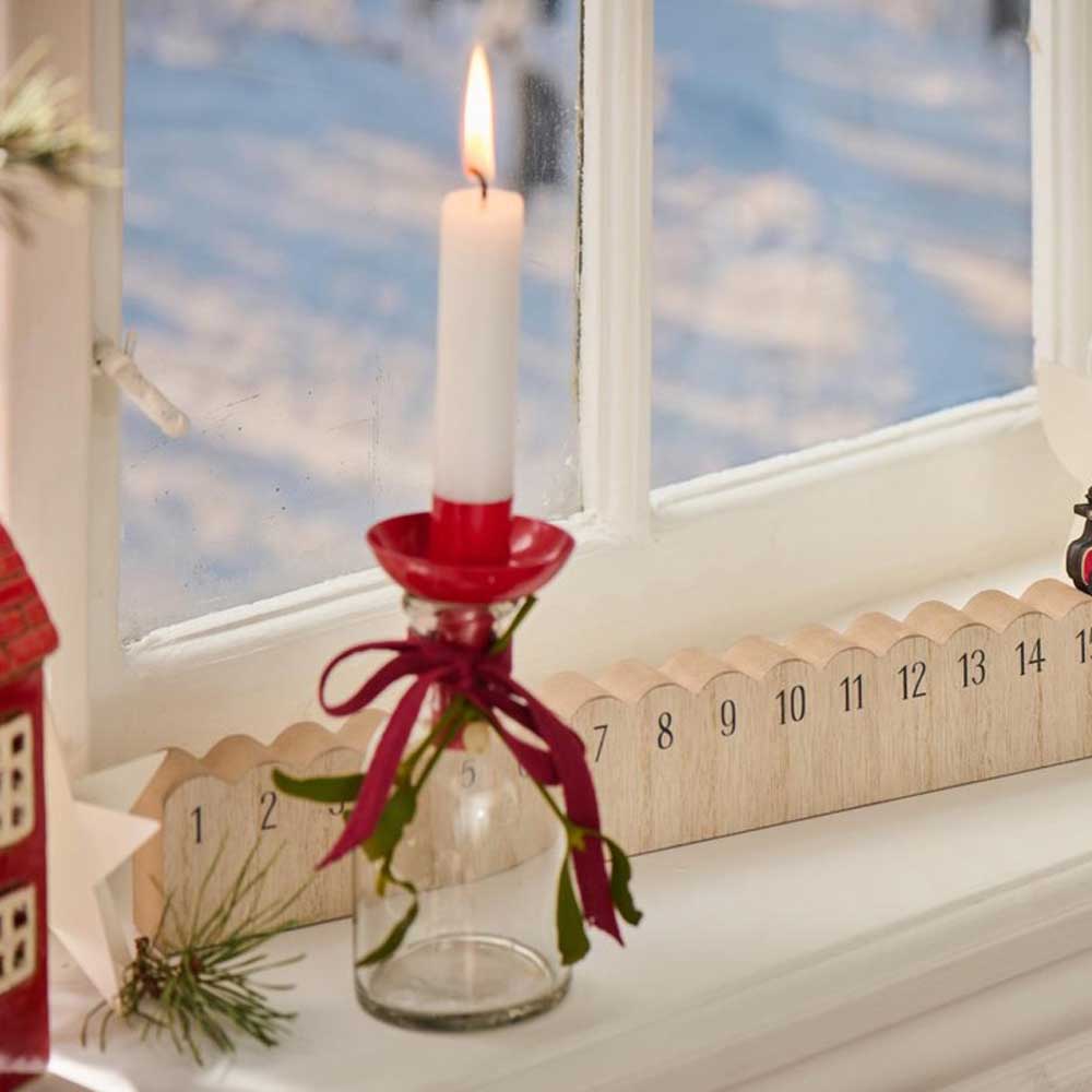 Auf einem Fensterbrett steht der elegant mit Grünpflanzen geschmückte Kerzenhalter von Ib Laursen, während eine brennende Kerze den roten Behälter neben einem nummerierten Holzkalender erhellt. Draußen ist die Szene mit Schnee bedeckt.