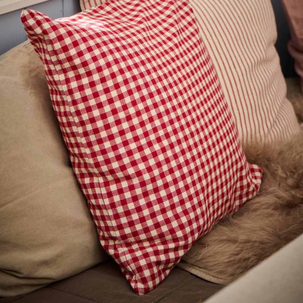 Ein roter Kissenbezug mit kleinen naturfarbenen Karos von Ib Laursen liegt neben einem cremefarbenen Streifenkissen auf einem beigen Sofa.