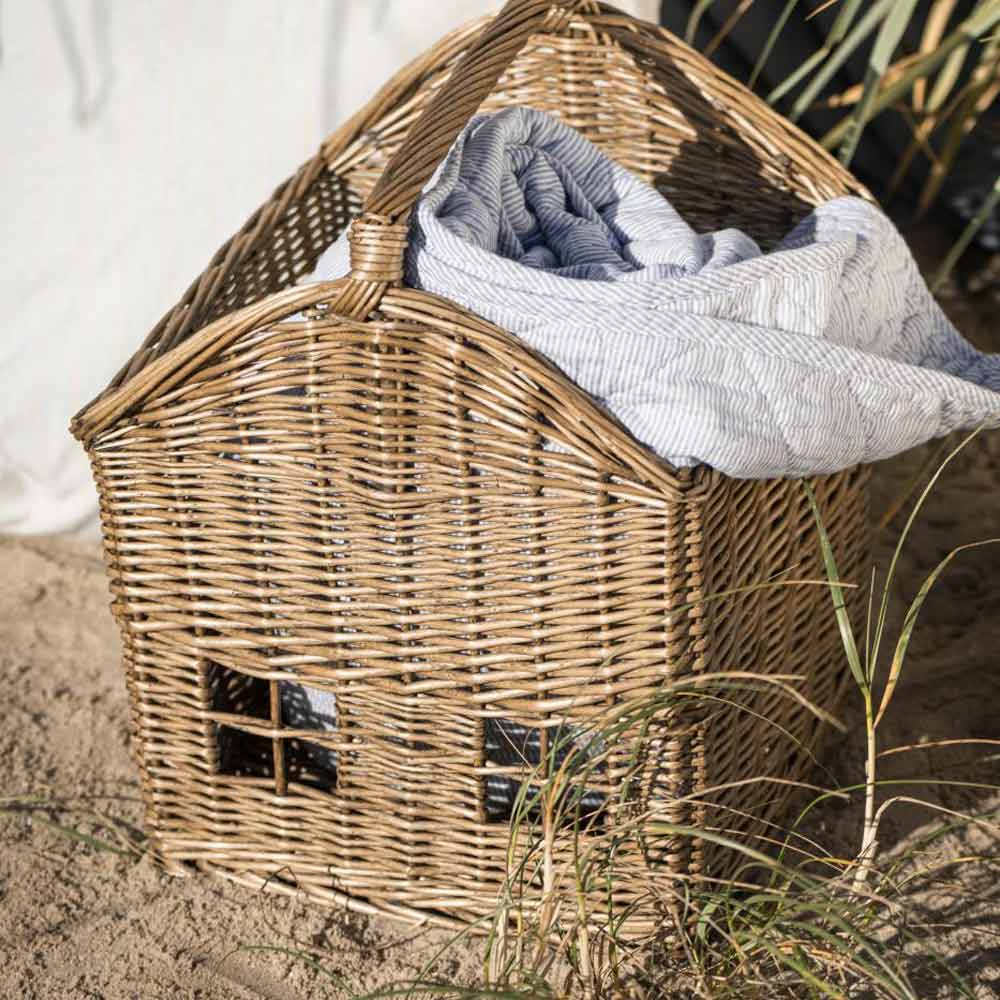 Ein Ib Laursen-Korbhaus in Form eines Hauses mit kleinen Fenstern und teilweise mit einer grau-weiß gestreiften Decke gefüllt, steht auf Sand neben etwas Gras.