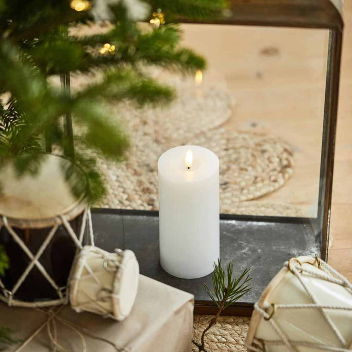 Eine Ib Laursen LED Stumpenkerze mit warmer Flamme steht auf einem dunklen Tablett, umgeben von verpackten Geschenken und kleinen Trommeln, in der Nähe liegt ein Tannenzweig.