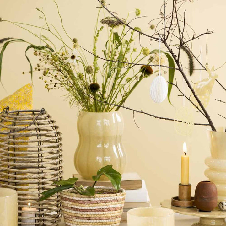 Ein ruhiges Stillleben-Arrangement mit einer Vase mit Wildblumen, dekorativen Zweigen, einer brennenden Kerze und einer Ib Laursen - Laterne Seegras und Metall mit Glaseinsatz auf beigem Hintergrund.
