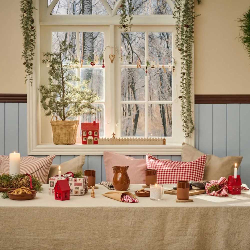 Eine gemütlich gedeckte Tafel mit dem Ib Laursen - Metallhaus Kerzenhalter für Tannenbaumkerzen in Rot, begleitet von Bechern, Tellern und Weihnachtsdekorationen vor einem mit Girlanden geschmückten Fenster mit Blick auf eine verschneite Winterlandschaft.