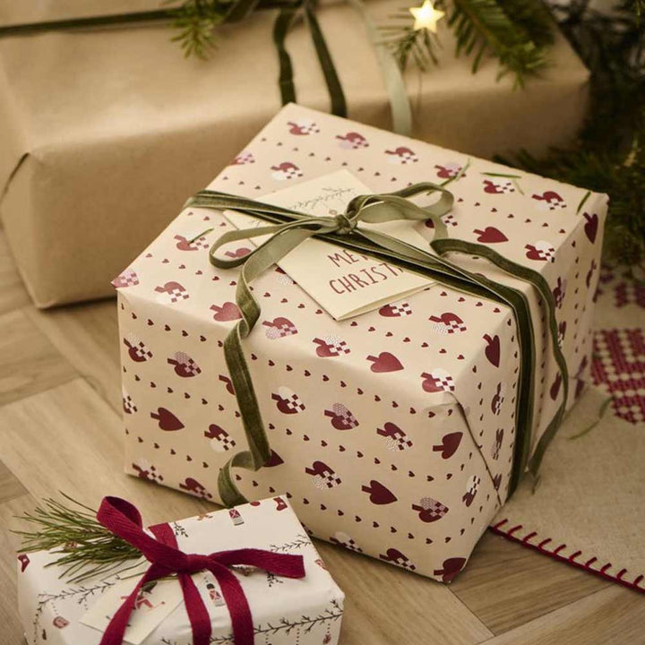 Zwei verpackte Weihnachtsgeschenke unter einem Weihnachtsbaum. Das größere Geschenk ist in festliche Ib Laursen Papierrolle Flechtherzen 5 Meter Rolle mit einem Etikett mit der Aufschrift „Frohe Weihnachten“ verpackt und das kleinere Geschenk ist mit einer roten Schleife verziert.