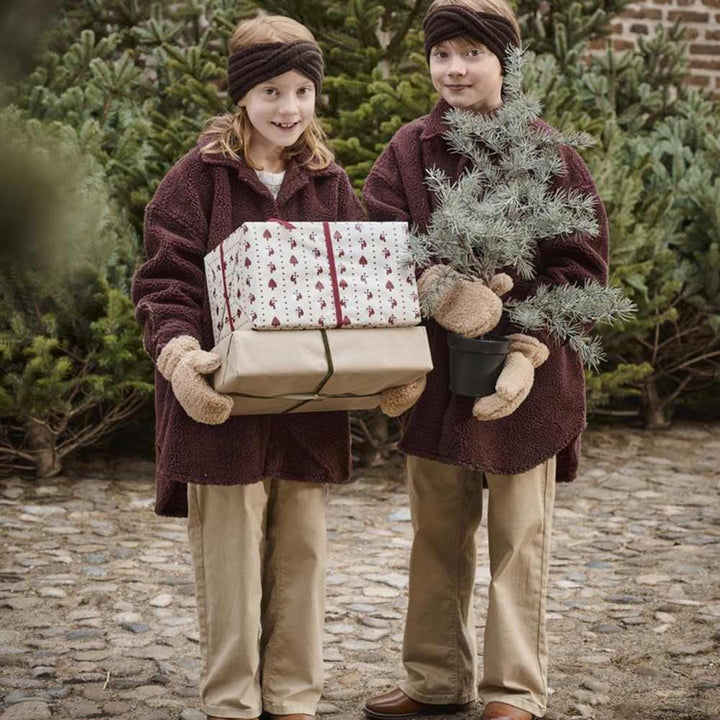 Zwei Kinder, warm gekleidet in braune Mäntel und Stirnbänder, stehen draußen und halten Geschenke und einen kleinen immergrünen Topfbaum in der Hand, der in 5 Meter Papierrolle Flechtherzen von Ib Laursen eingewickelt ist, umgeben von Grün.