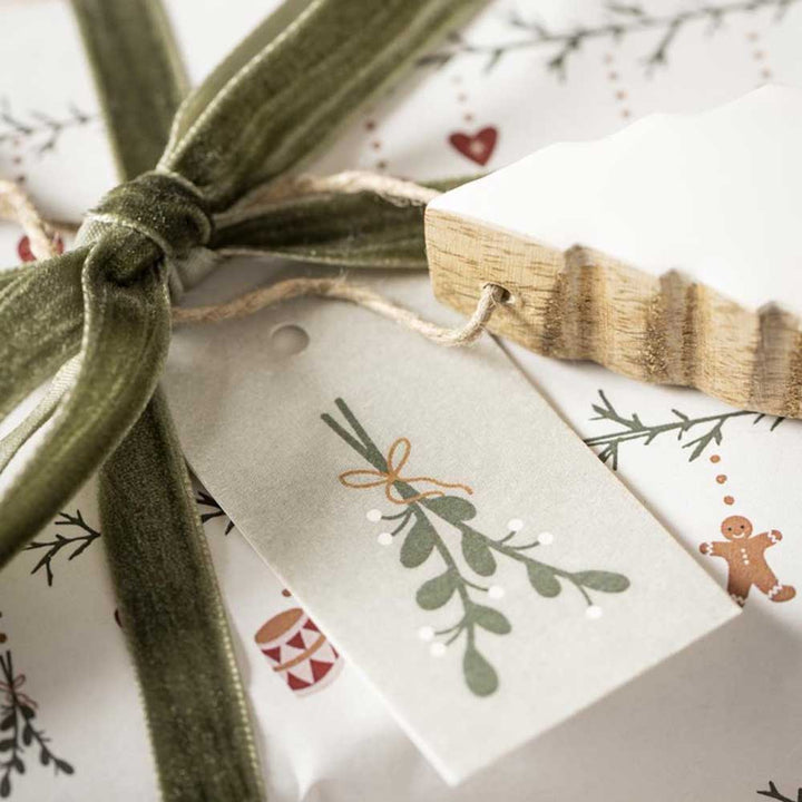 Eine Nahaufnahme eines verpackten Geschenks mit einer grünen Samtschleife, einem botanischen Geschenkanhänger und einem hölzernen Baumschmuck obendrauf. Das Geschenkpapier aus der Ib Laursen - Papierrolle My Nostalgic Christmas 5 mit Rolle-Kollektion weist ein festliches Muster auf.