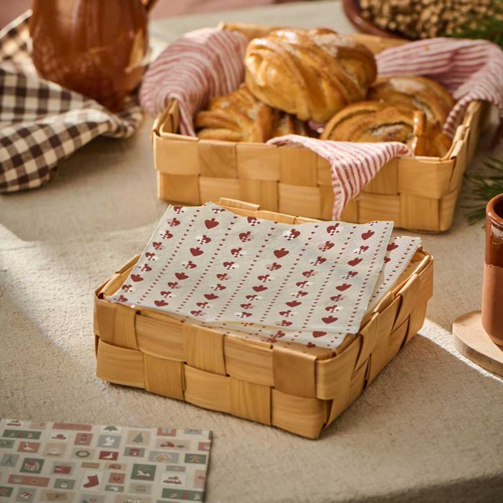 Ein Weidenkorb von Ib Laursen mit Gebäck steht auf einem Tisch neben einem weiteren Weidenkorb, der mit weißen Servietten mit dem Aufdruck „Papierserviette Flechtherzen“ gefüllt ist. Auf dem Tisch sind außerdem ein kariertes Tuch und ein brauner Becher zu sehen.