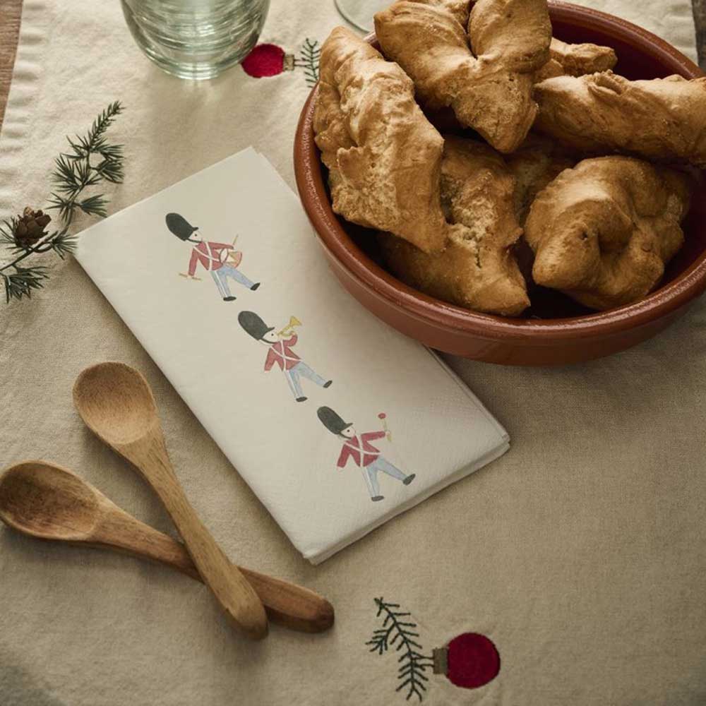 Eine Schüssel Brot, eine gefaltete Papierserviette „Gardisten“ von Ib Laursen mit Spielzeugsoldaten-Illustrationen, Holzlöffel und ein Glas auf einer bestickten Tischdecke.