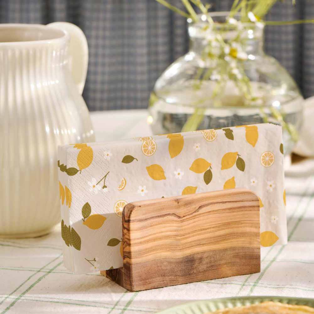 Auf dem Tisch steht ein Serviettenhalter Ib Laursen Papierserviette Zitrone (16 Stück), begleitet von einem weißen Keramikkrug und einer Glasvase mit Blumen.