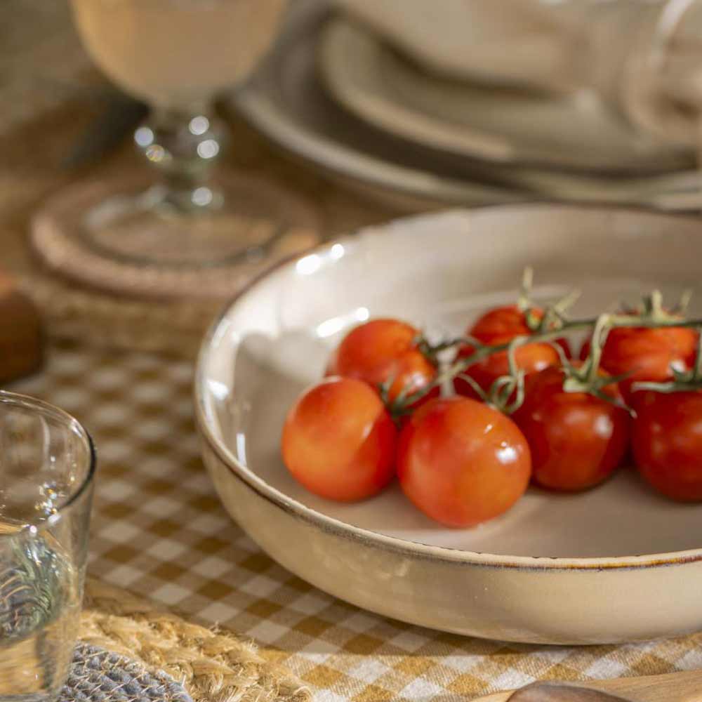 Eine Schüssel mit reifen Kirschtomaten auf einer karierten Tischdecke, mit einem Glas Wasser und einem Ib Laursen - Salat-/Pastateller Sand Dunes-Teller im Hintergrund.