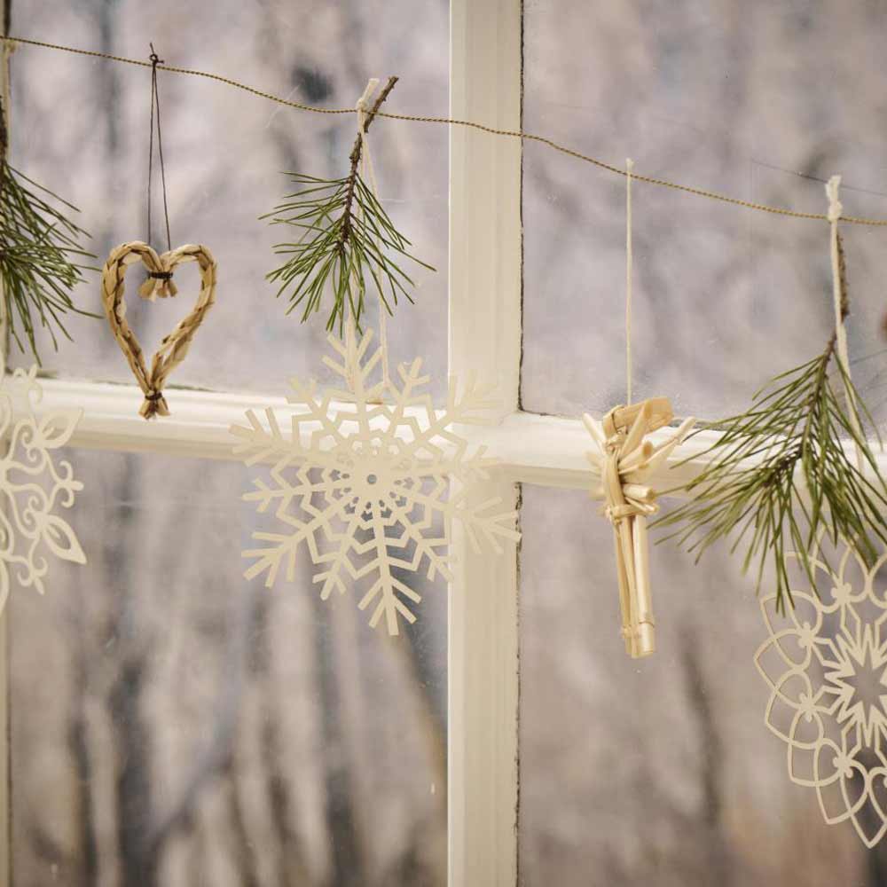 In einem Fenster ist eine mit hängenden Ornamenten geschmückte Girlande zu sehen, die ein Herz, mit Schnur umwickelte Tannenzweige und die Papierschneeflocken „Schneekristall“ von Ib Laursen Scherenschnitt umfasst und so eine winterliche Szene schafft.