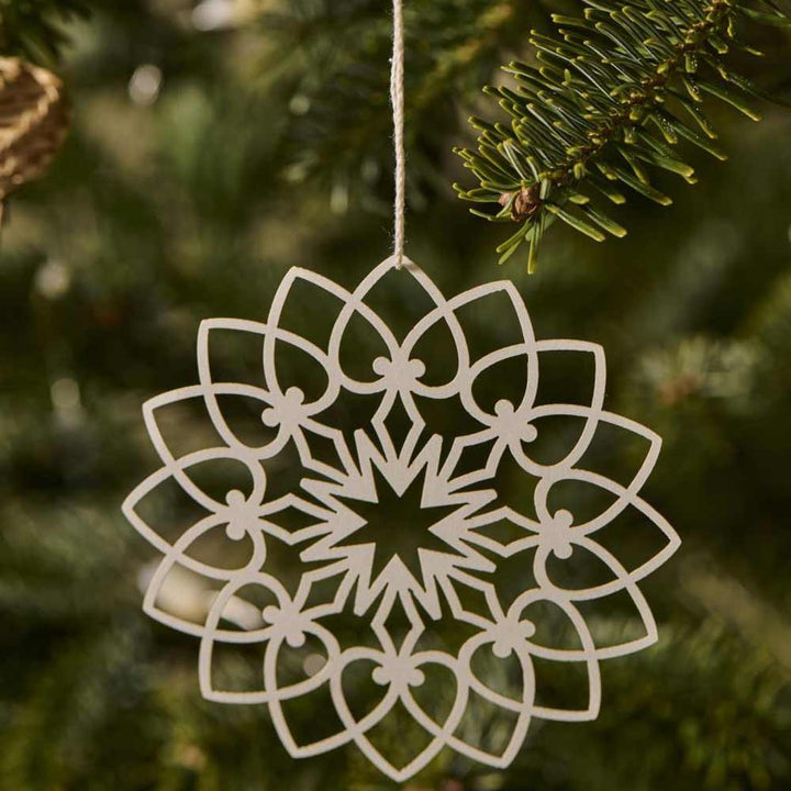 Ein aufwendig aus Holz gefertigtes Schneekristall-Ornament von Ib Laursen - Scherenschnitt hängt an einem grünen Tannenzweig.