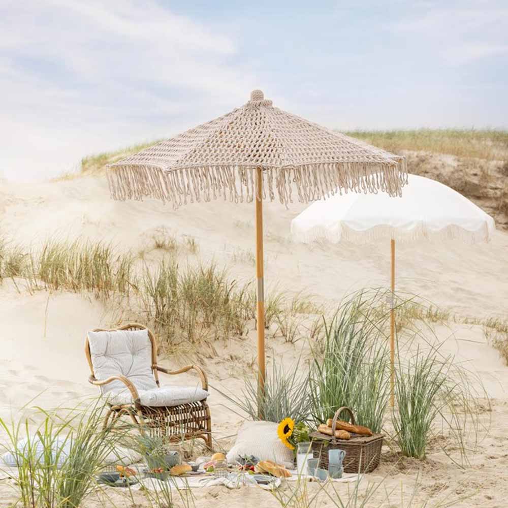 Eine Sandstrandszene mit zwei weißen Sonnenschirmen von Ib Laursen mit Fransen, einem Korbstuhl, einem Korb mit Gegenständen, einer Sonnenblume und viel Grün drumherum. Der Himmel ist blau mit wenigen Wolken und einige Dünengräser sind sichtbar.