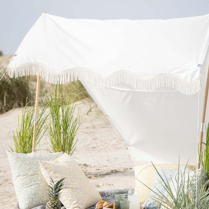 Eine Strandszene mit einem Ib Laursen - Sonnensegel weiß mit naturfarbigen Fransen, mehreren gemusterten Kissen und einer Ananas im Vordergrund. Im Hintergrund sind hohe Gräser und Sanddünen zu sehen.