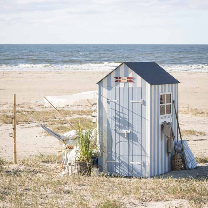 Eine kleine Strandhütte mit blau-weiß gestreiftem Design steht auf Sandrasen am Meer, daneben stehen ein Ib Laursen - Sonnensegel weiß mit naturfarbigen Fransen, ein Liegestuhl, ein Sonnenschirm und Strandaccessoires.