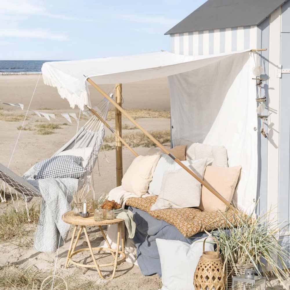 Eine gemütliche Strandausstattung mit einem Ib Laursen - Sonnensegel weiß mit naturfarbigen Fransen, Kissen, einer Hängematte und einem kleinen Tisch auf Sanddünen mit Blick auf eine ruhige Küste.