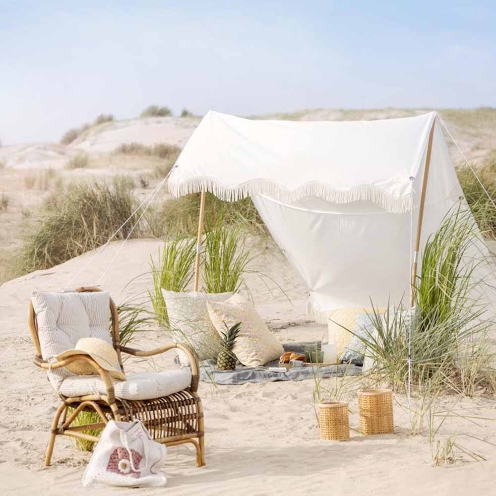 Ein kleines Ib Laursen - Sonnensegel weiß mit naturfarbigen Fransen, umgeben von Kissen, einem Korbstuhl und einem Picknick-Set, ist unter einem klaren Himmel auf den Sanddünen aufgebaut.