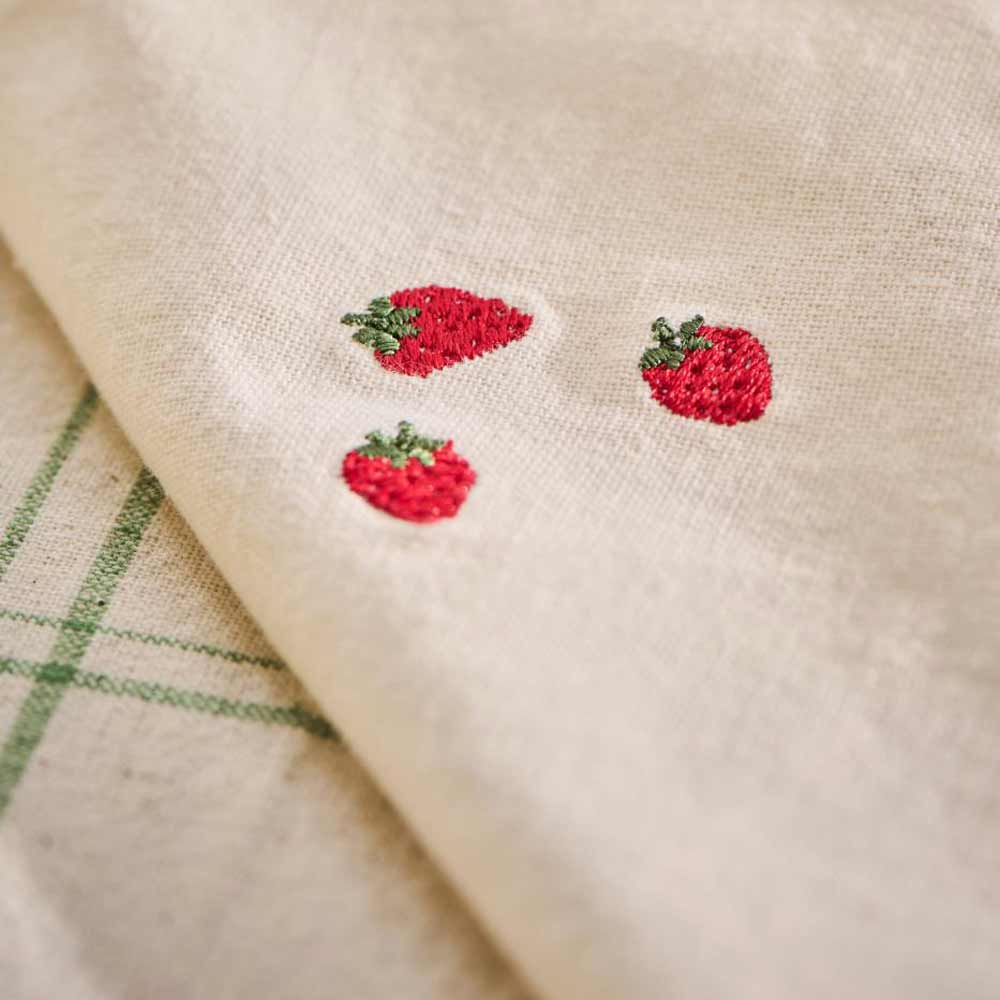 Nahaufnahme der „Stoffserviette Summer Vibes mit Erdbeeren Butter Cream“ von Ib Laursen, bestehend aus beigem Stoff mit drei kleinen aufgestickten roten Erdbeeren, die teilweise auf einem grün gestreiften Stoff platziert sind.