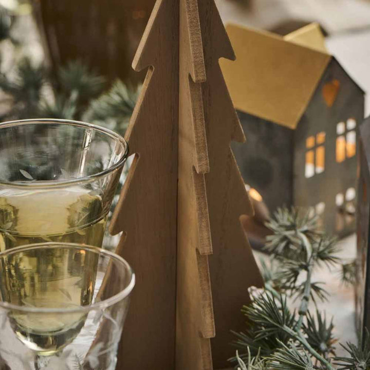 Zwei Gläser Weißwein neben einem Ib Laursen - Tannenbaum stehend aus Holz und kleinen beleuchteten Hausdekorationen, mit Tannenzweigen im Vordergrund.