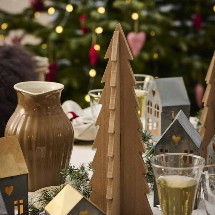 Das Foto zeigt einen Ib Laursen - Tannenbaum stehend Holz als hölzernen Weihnachtsbaumschmuck auf einem dekorierten Tisch, umgeben von kleinen hausförmigen Ornamenten, einem braunen Krug und Gläsern, mit einem beleuchteten Weihnachtsbaum im Hintergrund.