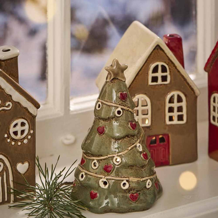 Ein mit Herzverzierungen verzierter Ib Laursen Tannenbaum stehend Stillenat steht vor Miniatur-Lebkuchenhäusern aus Keramik, eines davon mit einer roten Tür, auf einem Fensterbrett.