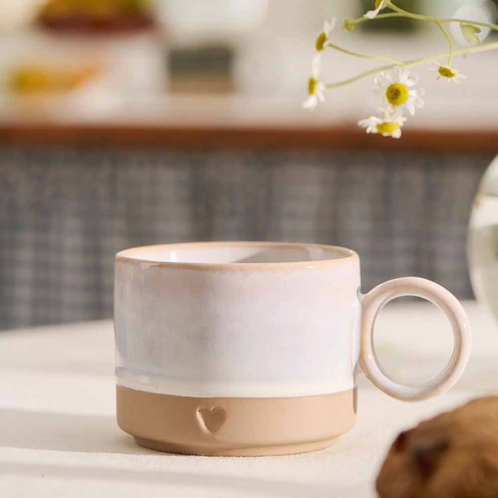 Die Ib Laursen - Tasse mit Henkel Herz mattbraunes Ton mit weißer Farbe steht auf einem Tisch neben weißen Gänseblümchen im Weichzeichner.