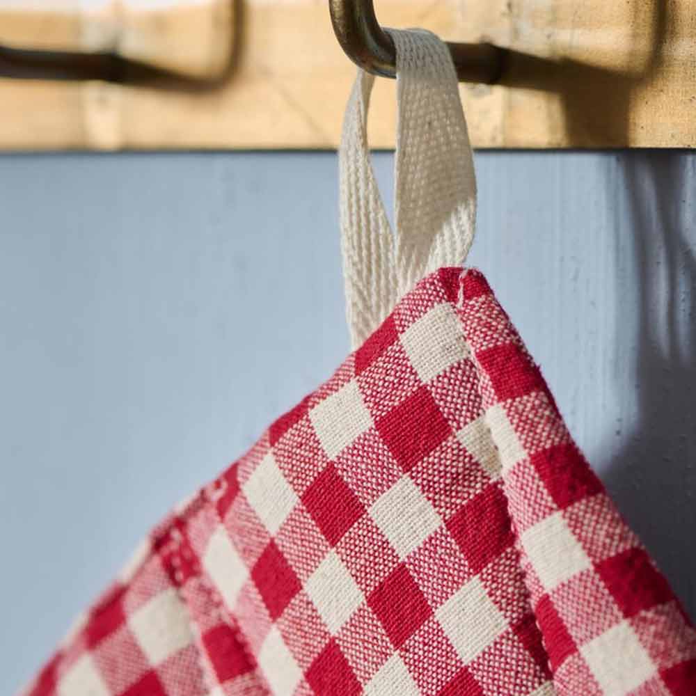 Nahaufnahme eines Ib Laursen - Topflappen Asta rot mit kleinen naturfarbigen Karos, der an einem Haken vor einer hellblauen Wand hängt.