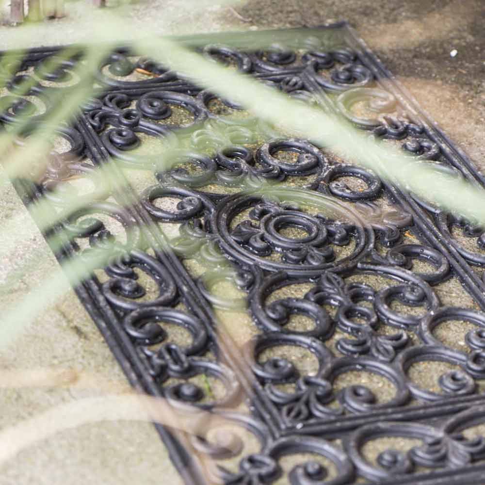 Dekorative Ib Laursen - Türmatte Gummi Filigran bedeckt mit Regentropfen auf einer sandigen Oberfläche, teilweise verdeckt durch verschwommenes grünes Laub im Vordergrund.